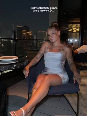 A woman sits in a chair on a balcony at night. She wears a light blue dress and has tattoos on her arms. The background shows city lights.