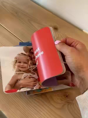 A woman holding a red book with the words 'just have!' printed on the cover. Inside the book are photos of a woman and a child. The woman is visible in the cover photo and is wearing a sun hat and holding the child. The hand of a person holding the red book is on the right side of the image, with a light colored wooden table in the background.