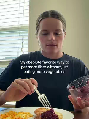 A medium shot shows a woman sitting at a table with food in front of her, holding a fork in her left hand and a small glass bowl with a jam-like substance in her right hand. The background includes a window with blinds. Text overlay at the bottom reads: "My absolute favorite way to get more fiber without just eating more vegetables". The composition features the woman at the center, a plate with food in the foreground, and a window with blinds behind her.