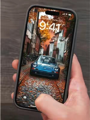 A hand holds a smartphone displaying a 4K wallpaper of a blue vintage car parked on a cobblestone street with autumn foliage, between European style buildings.
