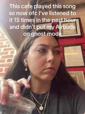 A young woman is sitting in a cafe with a drink in her hand and airpods in her ears, looking at the camera with a slightly annoyed or contemplative expression. The scene is lit with warm lighting and has frames and red wooden panels in the background.