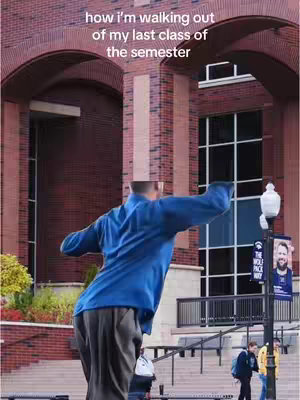 The image shows a man from the back with his arms raised, as though he's celebrating, walking out of a building with arched doorways. The building is made of brick, and the sky is overcast. The foreground includes stairs with various people walking by.