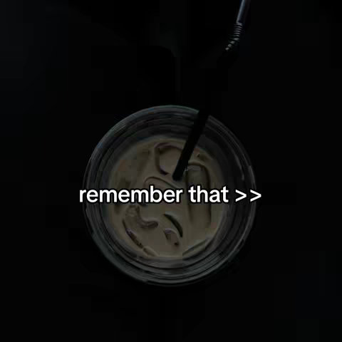 A top-down shot of a clear glass filled with a creamy, brown liquid. A black straw is inserted into the drink. The background is a solid black.