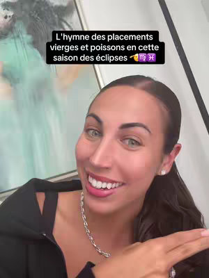 A woman with dark hair and a necklace is smiling and pointing to the right. The background is blurry, with some artwork visible. A text overlay reads, 'L'hymne des placements vierges et poissons en cette saison des éclipses'.