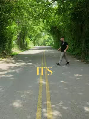 A man walks down a road flanked by lush green trees, their foliage forming an arch over the road. The text 'IT'S' is superimposed in a yellow font on the road surface.