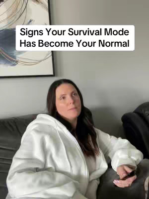 A woman sits on a couch in what appears to be a home setting. She's wearing a white hoodie and looking off to the side with a thoughtful expression. Above her head is a white rectangle with the text 'Signs Your Survival Mode Has Become Your Normal'. The background includes a framed piece of art and a section of the couch.