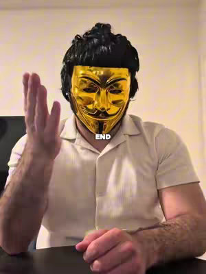 A man wearing a gold Guy Fawkes mask and a light tan shirt, raising his hand with what appears to be a stick for an extended arm. The background is blurred and interior.