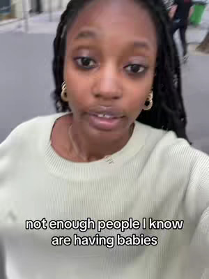 A young Black woman stands outdoors, facing the camera. Her dark, braided hair is pulled back, and she wears gold hoop earrings and a light green sweater. A text overlay at the bottom reads, "not enough people I know are having babies."