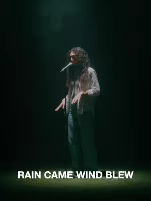 The image features a person standing in a dark setting, lit by a spotlight from above. The person is holding a microphone, and text at the bottom of the image reads "RAIN CAME WIND BLEW."