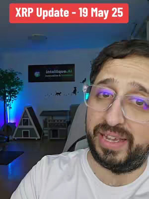 A man with glasses is centered in the frame, smiling and speaking. He is wearing a white t-shirt. Behind him, the background includes a wall with a logo that reads 'intelliique.ai'. There's a cabinet on the left side, and a modern white cabin-shaped object beside the cabinet. The lighting seems to be a mix of natural light and artificial blue light.