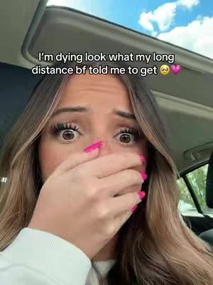 A young woman with long, light brown hair and bright pink nail polish is covering her mouth with her right hand. Her eyes are wide with a look of surprise or shock. The background suggests she is inside a car with a bright sky visible through the windshield.