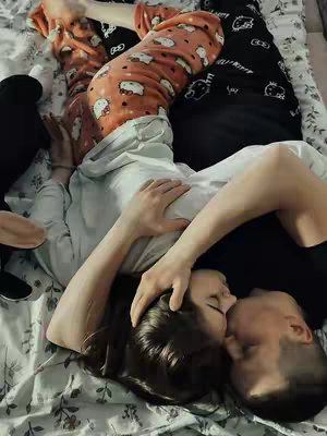 A couple lying on a bed embracing and kissing. The woman is on the bottom and the man is on top kissing her. The bed has a floral pattern. The woman is wearing a light gray shirt with her hair down. The man is wearing a black shirt and the bodies of a third person are on each side of the frame in the background wearing pajamas. The lighting is natural.