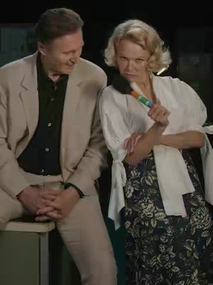 A still from a film featuring Liam Neeson and Pamela Anderson. Liam Neeson, on the left, is wearing a tan suit. Pamela Anderson, on the right, is holding a colorful microphone. The background appears to be a set.