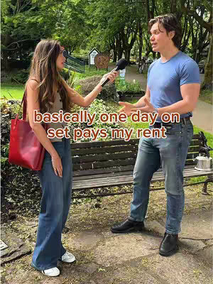 A woman interviews a man outdoors. The woman, on the left, holds a microphone and red tote bag. The man, on the right, is mid-sentence, gesturing with his hands. Behind them are benches and foliage.