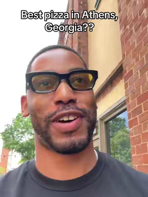 A man with short hair and a beard wears black-framed glasses and a black t-shirt. The text overlay reads "Best pizza in Athens, Georgia??" The background includes a brick building and part of a window.