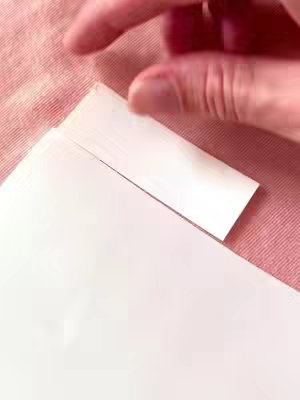 A close-up shot of a person's hand gently lifting a corner of a stack of white paper, revealing a clean, lined edge. The background is a soft, textured pink surface. The lighting appears to be natural, creating a soft, even tone.