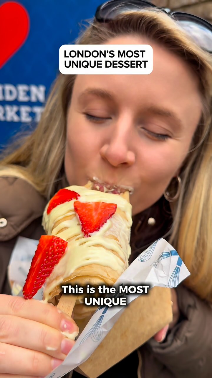 A woman is shown taking a bite of a dessert, which appears to be a crepe wrapped around a Magnum ice cream bar. It's drizzled with white chocolate and topped with fresh strawberries. The image is framed from a close-up angle, focusing on the woman and the dessert. Text overlays add information about the dessert, while the background is slightly blurred.