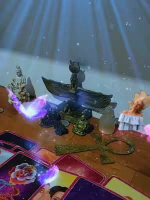 The cover image shows a collection of tarot cards and spiritual objects arranged on a wooden surface, likely for a tarot reading. Various items such as statues, ankh, and other symbolic items are also placed around the tarot cards. The overall composition is top-down with a blue and purple ethereal glow.