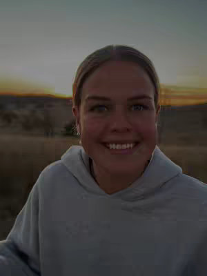 A woman wearing a gray hoodie smiles at the camera in front of a dusky, golden-hued landscape. The sky transitions from warm oranges and yellows to a hazy, indistinct top, likely representing either sunrise or sunset. The woman's face is centered, with her expression conveying warmth and friendliness. The background suggests an outdoor setting near hills or a natural area.