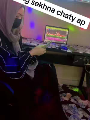A woman wearing a hijab and a mask in front of a desk covered in money stands next to a laptop displaying a trading chart, with colorful lights in the background.
