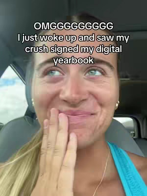 A close-up shot of a young woman in a car. She is smiling, with her hand covering her mouth, suggesting a surprised or excited expression. Text overlays on the image read: "OMGGGGGGGGG I just woke up and saw my crush signed my digital yearbook". The background includes car details like a seat and dashboard.