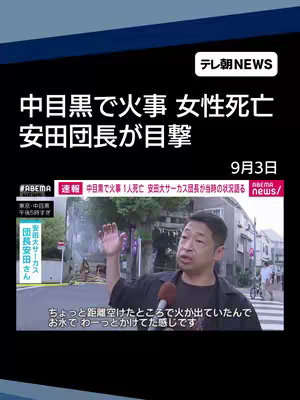 A news report cover image featuring a man speaking into a microphone. The setting appears to be near a street with buildings and green vegetation. Above, a news headline announces a fire incident in Nakameguro and a woman's death. The 'ABEMA news' logo is visible, as is the 'TV Asahi News' logo. The man is gesturing with his right hand, and the image is framed with a dark blue upper and lower border.