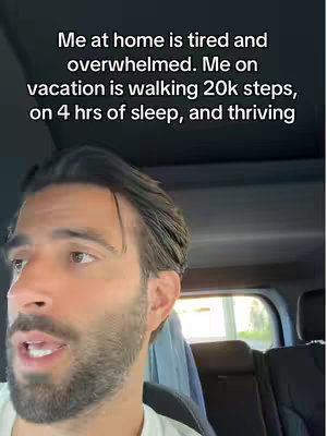 A man is filming himself from inside a car. Text overlays state his energy and activity levels at home versus on vacation. The man has short, dark hair and a well-groomed beard. He appears to be looking and talking at the camera. The overhead lighting creates a shine on his hair.