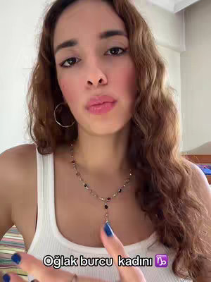 Image shows a young woman from the chest up, gazing at the camera. She has long, wavy brown hair, large hoop earrings, and is wearing a white tank top and a necklace. Text at the bottom reads 'Oğlak burcu kadını ♑'. The background is blurred, and the woman is the focus of the image.