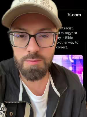 A man with glasses and a beard faces the camera head-on, wearing a baseball cap and a black jacket with a white t-shirt underneath, while a phone screen displaying text is in the background.