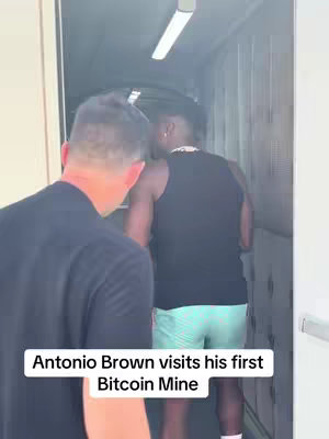 Two men are walking down a narrow hallway or corridor. The person in the foreground is wearing a black t-shirt, and the back of the person is visible. The person in the background is wearing a black tank top and green shorts.