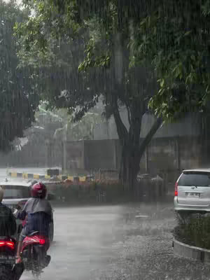 A street scene in a heavy rainstorm. A person on a motorcycle rides down a wet road, with blurred buildings and trees in the background. The sky is grey and filled with falling rain, creating a sense of being in the rain. The composition is centered around the bike and the road.