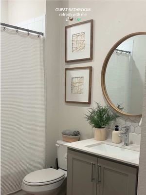 A well-lit photo of a clean and organized guest bathroom, showcasing a white and gray color scheme with a touch of greenery. The bathroom features a white shower curtain and a gray vanity with a white countertop and a silver faucet. A round mirror hangs above the sink, and two framed pictures are mounted on the wall. A small potted plant adds a natural element to the space. The overall composition is neat and inviting.
