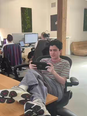 A young man sits with their legs on a desk, playing a handheld gaming device. They are in an office setting with other people in the background. The composition is casual and modern, with a focus on the individual gaming.