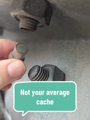 Close-up shot of a hand holding a cylindrical object resembling a key or capsule. The object is placed near two large bolts and nuts, one of which has been partially unscrewed. A speech bubble with text appears at the bottom.