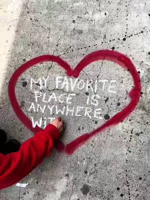 A close-up shot of a gray concrete wall with a large red heart spray-painted on it. Inside the heart, white chalk writing says "MY FAVORITE PLACE IS ANYWHERE WITH." A person's arm, wearing a red sleeve of a jacket, is seen writing with chalk. Black paint splatters are randomly distributed around the heart on the wall.