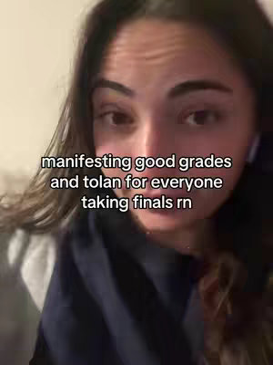 A close-up shot shows a young woman with long, dark hair looking directly at the camera. Text overlayed on the image reads, “manifesting good grades and tolan for everyone taking finals rn.” The background is softly lit, suggesting an interior setting.