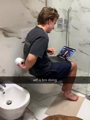 A man is sitting on a toilet in a bathroom, holding a roll of toilet paper and looking at a laptop. The laptop screen displays a video of two people. The bathroom has a modern design with white marble-like walls. A water bottle is visible near the laptop.