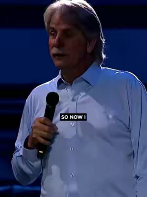 A man with graying hair and a mustache is speaking while holding a microphone. He is wearing a light blue button-down shirt. The background is a dark blue.