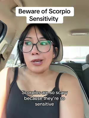 A selfie-style shot taken inside a car. A woman with dark hair and glasses is the primary focus, sitting in the driver's seat. Above her head is a white rectangular text overlay that reads: "Beware of Scorpio Sensitivity." Below, a similar text overlay states: "Scorpios are so scary because they're so sensitive." The background suggests a sunny outdoor setting.