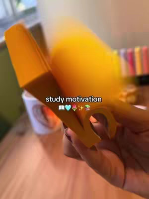 The image shows a hand holding a large yellow object, possibly a highlighter, up towards the camera. The background is blurred, but it appears to be a desk or table with a white mug and some colorful stationary supplies such as pens. Overlayed on the image is the text 'study motivation'.