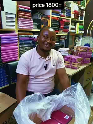 A man with short hair smiles warmly, seated in a shop filled with bookshelves. He's wearing a light pink polo shirt and holding a white bag with red items inside. Bookshelves are stacked with books in various colors, adding depth and a commercial setting.