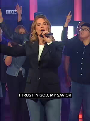 The cover image shows a well-lit stage with a woman in a dark blazer and blue jeans holding a microphone and singing with other people in the background. The background contains colorful lights.