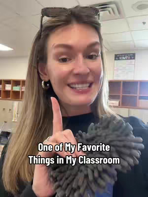 A woman with glasses on her head is pointing her right index finger upwards. The image is taken in a brightly lit classroom setting.