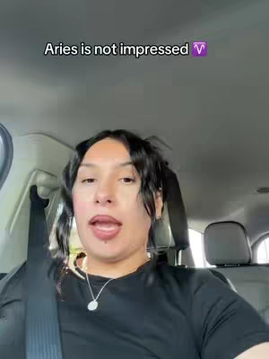 A woman is filmed from the front, sitting in the driver's seat of a car. She has dark hair pulled back into two small pigtails and is wearing a black top and a necklace. The car interior is visible, with headrests and seatbelts. Text overlay at the top says Aries is not impressed.