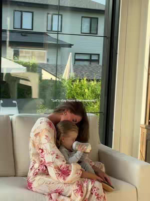 A woman and a young child are seated on a cream-colored sofa. The woman wears a floral dress and embraces the child, who is drinking from a bottle. The backdrop reveals a large window with a view of a house, a yard, and the sunny sky. The overall composition conveys a sense of warmth and domestic tranquility.
