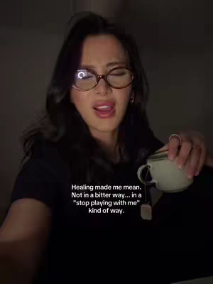 A woman with dark hair and glasses is mid-sentence, holding a teacup and looking slightly up and to the side.