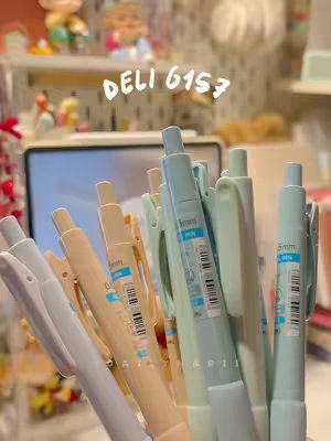 Close-up shot of a collection of colorful pens. The pens are primarily light blue, light green, and beige, with their tips pointing up towards the viewer. The background is softly blurred, with a hint of a shelf with small decorations and a tablet.
