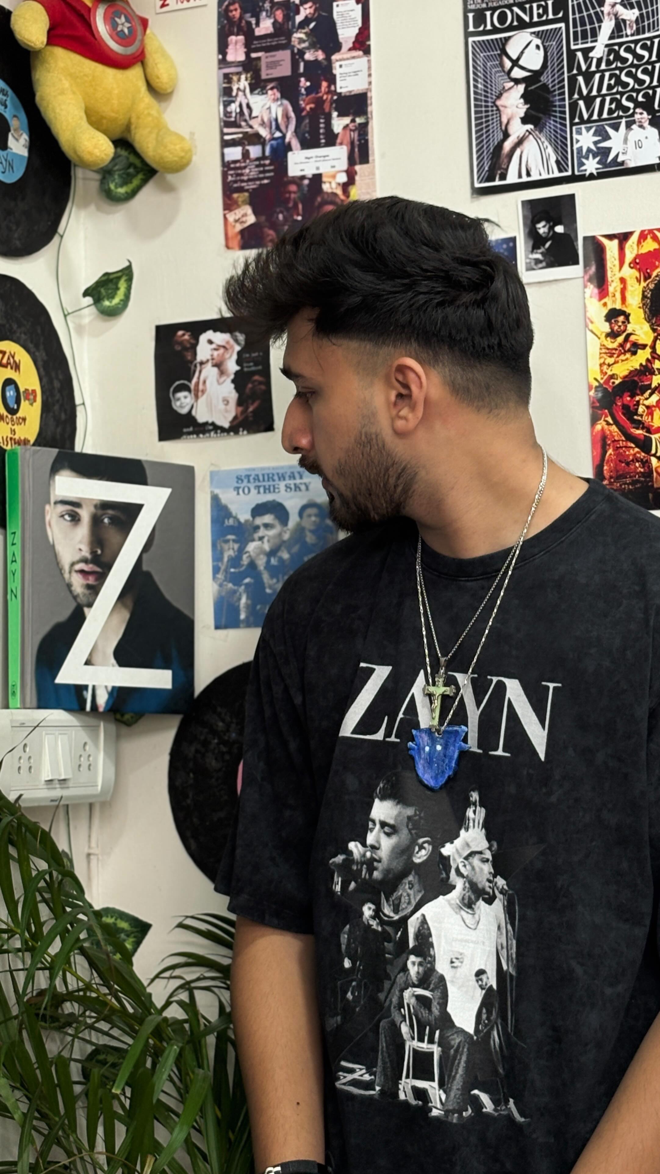 A man with dark hair and a trimmed beard stands angled slightly to the left, looking over the shoulder. The background shows a wall decorated with several framed Zayn Malik album covers, posters, and vinyl records. He is wearing a black t-shirt with a graphic of Zayn Malik, accessorized with silver necklaces featuring a pendant. The lighting seems to be natural, highlighting the man's face and the details of his outfit and the room. There's a plant in front of him.