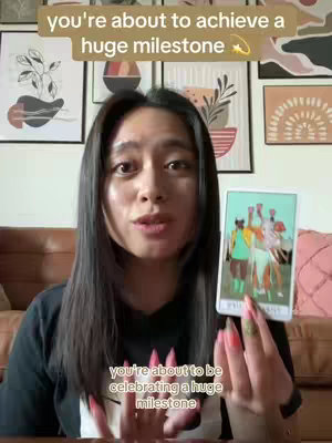A woman is centered in the frame, holding a tarot card. She has long, dark hair and is looking directly at the camera. The background shows various framed pictures on the wall, and a couch with a decorative pillow.