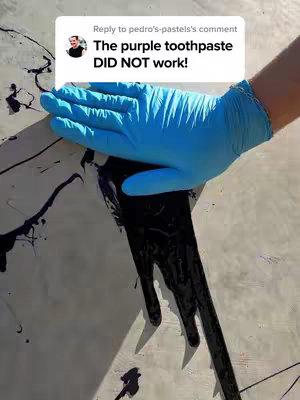 A photograph shows half of a person's body interacting with black, viscous liquid on a lightly colored surface.  A hand covered by a bright blue glove presses down into the liquid near the center of the image. Dark streaks of the liquid run from the hand down the surface. A speech bubble, white with black text, hovers in the top left: "The purple toothpaste DID NOT work!" A small image of a person in a black shirt is present in the speech bubble.